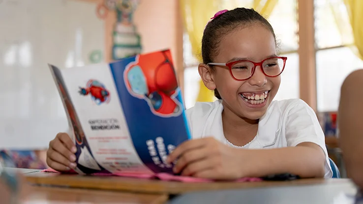 Smiling girl in red glasses holding a colourful Superbook booklet in a classroom setting