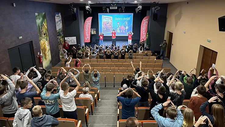 Children in an auditorium enthusiastically participating in a Superbook show, with presenters on stage and a vibrant screen display
